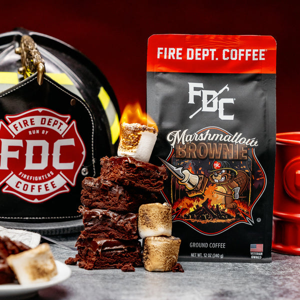 A bag of Fire Department Coffee's 12 oz Marshmallow Brownie Coffee with a red section accross the top that says "Fire Dept. Coffee". The art on the bag features a beaver holding a pike pole with a marshmallow and brownie on it over a campfire. The title reads, "Marshmallow Brownie" and there is a veteran owned badge with an American flag in the bottom right corner.