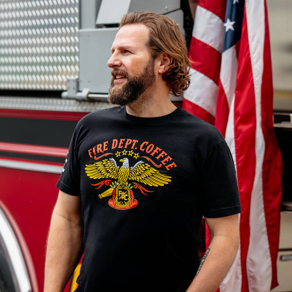 The front side of a black t shirt. On the chest of the t shirt is an eagle holding an FDC fire helmet. Above the eagle is text that reads "Fire Dept. Coffee"